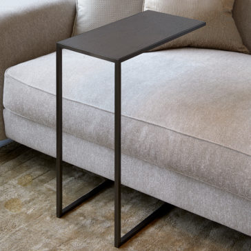 Contemporary Designer Over Sofa Side Table The Contemporary Designer Over Sofa Side Table, featuring a slender black metal frame and a dark wooden top, is elegantly situated on a beige textured rug. It sits seamlessly next to a light grey upholstered sofa with luxurious backrest and cushions, perfecting the modern, minimalist aesthetic.