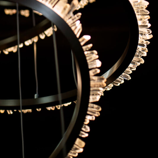Modern Quartz Crystal Hoop Chandelier Close-up image of the Modern Quartz Crystal Hoop Chandelier featuring circular frames adorned with crystal-like elements. The black metal rings are beautifully illuminated by lights, highlighting intricate details and creating a luxurious, elegant ambiance against a dark background.