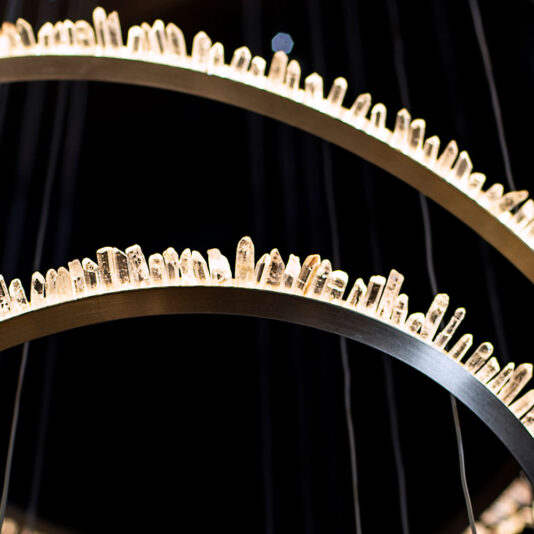 Modern Quartz Crystal Hoop Chandelier A close-up view of the Modern Quartz Crystal Hoop Chandelier showcases its circular metal rings adorned with upright crystal-like rods. These rods emit a soft, warm glow against a dark background, creating an elegant and contemporary aesthetic.