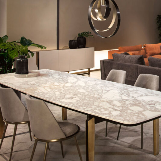 Modern Rectangular Marble Dining Table A modern dining room showcasing the Modern Rectangular Marble Dining Table with gold legs, surrounded by beige upholstered chairs. In the background, contemporary wall art can be seen along with a beige sideboard, indoor plants, and a brown sectional sofa. The space is well-lit and stylishly decorated.