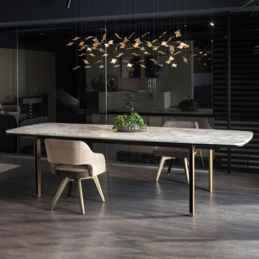 A modern dining room featuring the Modern Rectangular Marble Dining Table with gold legs and two cushioned chairs. A centerpiece of green foliage sits on the table. Above, a chandelier with numerous hanging, golden geometric shapes adds an artistic touch to the sleek space.