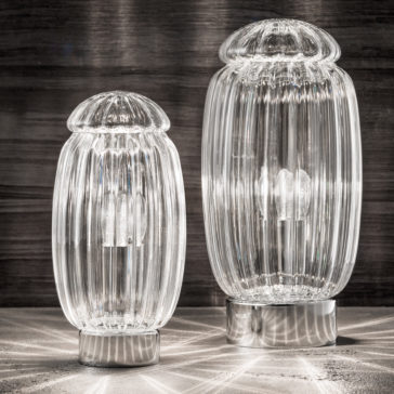 Two Modern Silver Murano Glass Table Lamps with ribbed cylindrical bodies and dome-shaped tops sit on a reflective surface. The larger lamp, positioned on the right, casts intricate light patterns onto the surface, while the smaller lamp, on the left, does the same.