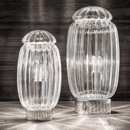 Two Modern Silver Murano Glass Table Lamps with ribbed cylindrical bodies and dome-shaped tops sit on a reflective surface. The larger lamp, positioned on the right, casts intricate light patterns onto the surface, while the smaller lamp, on the left, does the same.