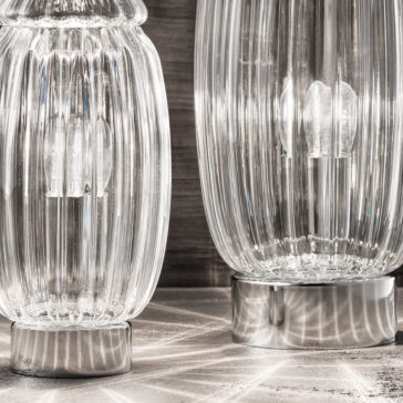 Close-up of two elegant, ribbed glass table lamps with a wavy texture, emitting light. The lamps have metallic bases and cast intricate shadows on the surface beneath them. In the background, a textured, dark wooden panel complements the Modern Curved Sofa With Brass Frame.