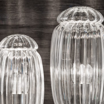 Close-up of two decorative glass light fixtures with ribbed translucent shades. The lights are turned on, emitting a soft white glow. The Modern Curved Sofa With Brass Frame resides in the blurred background, enhancing the detail and elegance of the glass design.