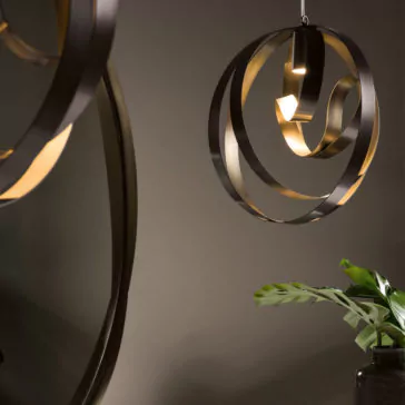 A Modern Twisted Bronze Chandelier featuring artistic, ribbon-like, circular black and gold elements is suspended next to a dark round mirror. Below the fixture, a green plant with broad leaves adds a touch of nature to the sophisticated decor as it rests on a surface.
