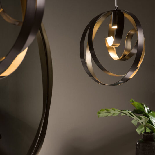 Modern Twisted Bronze Chandelier A Modern Twisted Bronze Chandelier featuring artistic, ribbon-like, circular black and gold elements is suspended next to a dark round mirror. Below the fixture, a green plant with broad leaves adds a touch of nature to the sophisticated decor as it rests on a surface.