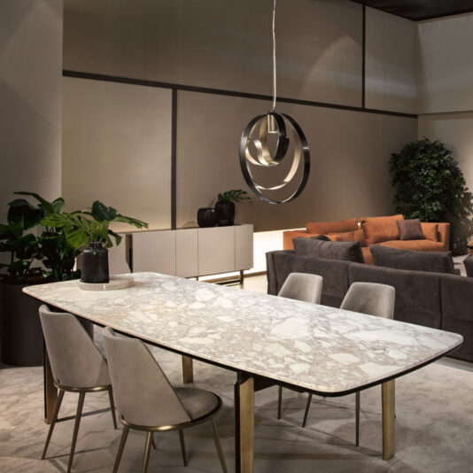 Modern Twisted Bronze Chandelier A contemporary living and dining room highlights a long, marble-top dining table paired with four gray upholstered chairs. A Modern Twisted Bronze Chandelier is suspended above the table, adding a unique touch to the space. In the background, an orange sofa, gray armchairs, a sideboard, and potted plants complete the stylish decor.