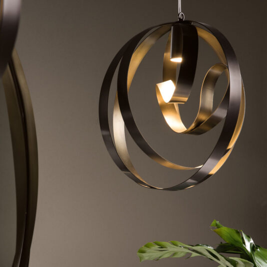 The Modern Twisted Bronze Chandelier, featuring swirling metallic ribbons in both dark and golden hues, hangs from the ceiling. Its stylish design creates a sophisticated ambiance. A green plant is partially visible in the lower corner, adding a touch of nature to the scene.
