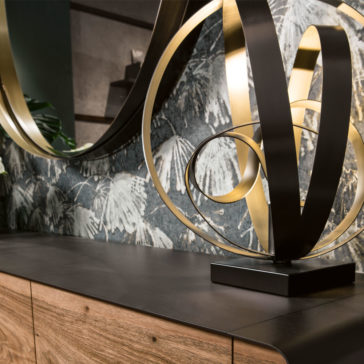 Modern Twisted Bronze Table Lamp A chic and modern interior is showcased with a close-up of a black countertop featuring a sleek gold and black abstract sculpture. Beside it, the **Modern Twisted Bronze Table Lamp** adds an artistic touch. The countertop sits atop a wooden cabinet, with leafy patterned wallpaper and a large, stylish mirror in the background.