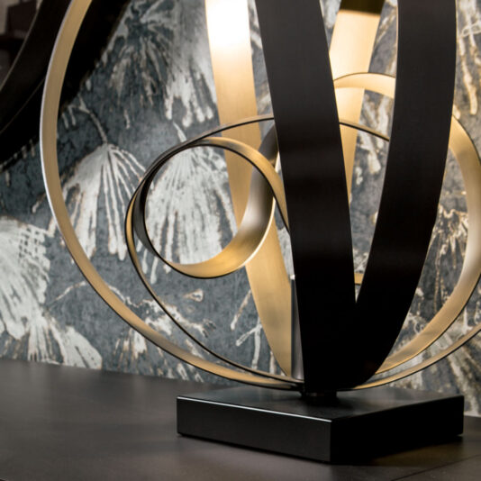 Modern Twisted Bronze Table Lamp A close-up of a sculpture characterized by intertwining black and gold metallic ribbons on a black base, evocative of the Modern Twisted Bronze Table Lamp. The backdrop showcases a textured surface adorned with a monochromatic feather-like pattern.