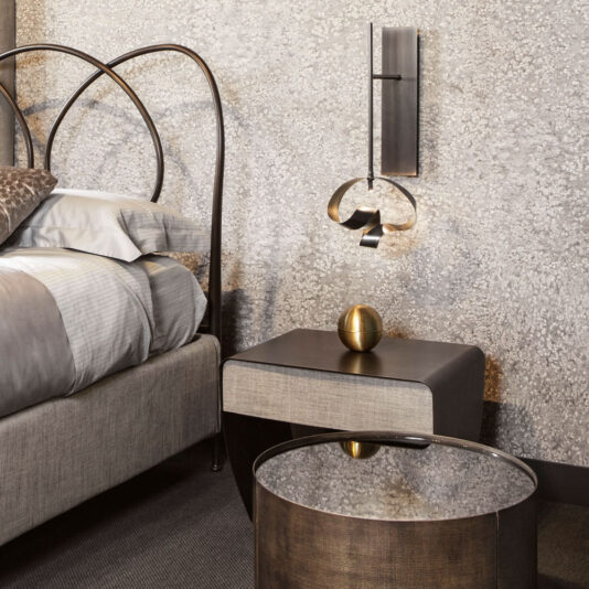 A modern bedroom showcasing a bed with a curved metal headboard and neutral-toned bedding. A sleek nightstand supports a gold spherical sculpture, while above it hangs an abstract wall-mounted light fixture, complemented by the Modern Twisted Bronze Wall Light. The walls feature a textured finish.