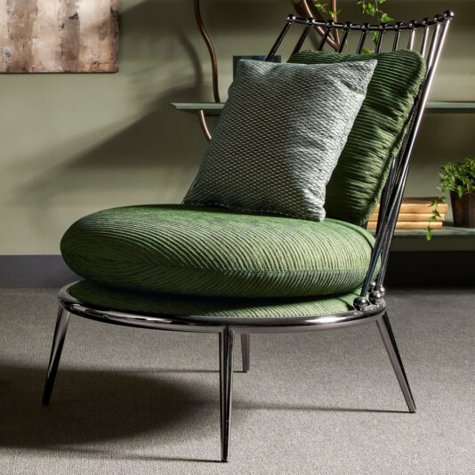 The Modern Upholstered Iron Occasional Chair features a shiny metal frame and four slender legs, with a round, green, textured seat cushion and a matching back cushion. A green pillow rests against the back cushion. This stylish chair is set against a neutral wall and carpeted floor.