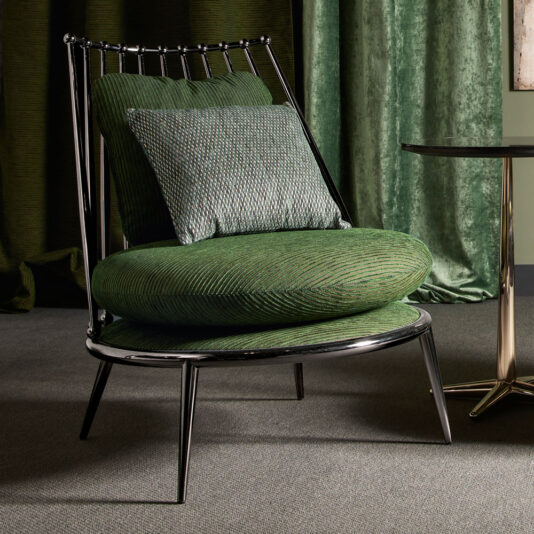 Modern Upholstered Iron Occasional Chair A Modern Upholstered Iron Occasional Chair with green fabric sits beside a small, round black table supported by a single pedestal base. The chair features both a circular seat and backrest, each adorned with matching green cushions. In the background, there is a green curtain hanging.
