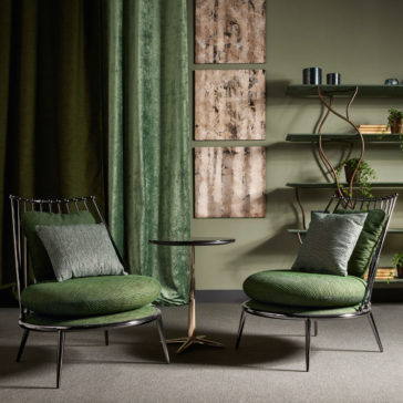 A cozy sitting area features two Modern Upholstered Iron Occasional Chairs, each adorned with accent pillows, surrounding a small round side table. Behind the chairs, green drapes frame three vertically hung abstract wall art pieces, alongside a modern shelf decorated with plants and various decor items.