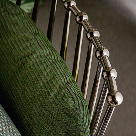 Modern Upholstered Iron Occasional Chair Up-close view of the Modern Upholstered Iron Occasional Chair showcasing a polished chrome backrest adorned with spherical caps. The iron frame gracefully supports a textured green cushion with a wavy pattern, striking a stylish contrast between the metallic elements and the fabric.