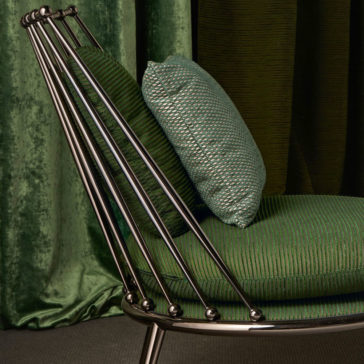 The Modern Upholstered Iron Occasional Chair, showcasing a sleek metallic iron frame and green cushions, is positioned against a backdrop of dark green curtains. This stylish chair features vertical metal rods for back support, with the cushion displaying a textured, striped pattern and an extra green pillow added for comfort.