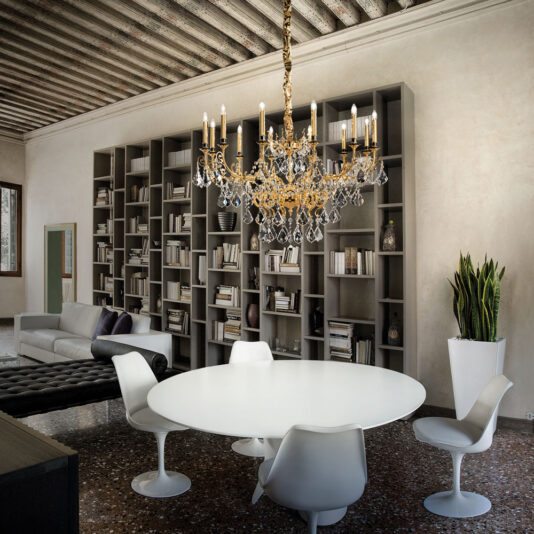 A contemporary living area features a large bookshelf filled with books, a gray sectional sofa, a black tufted ottoman, and a white round dining table with four modern white chairs. The Opulent Crystal Pendant Chandelier hangs from the wooden beamed ceiling above the table.