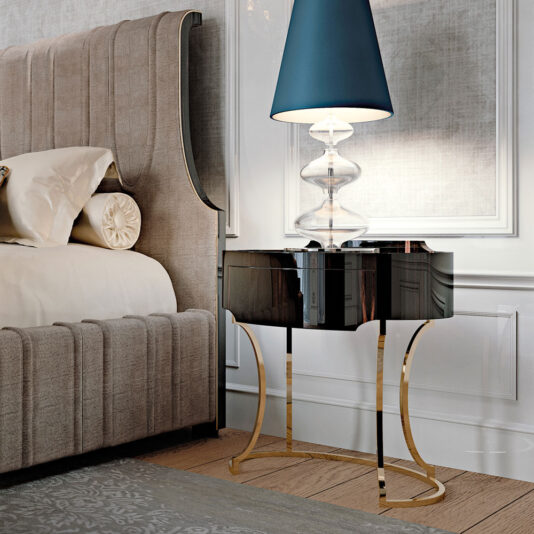 A stylish bedroom corner features a beige upholstered bed with a decorative pillow and bolster. An Oval Italian Ebony Bedside Table with golden curved legs holds a modern glass lamp with a blue shade. The room's decor is elegant and contemporary, with soft lighting.