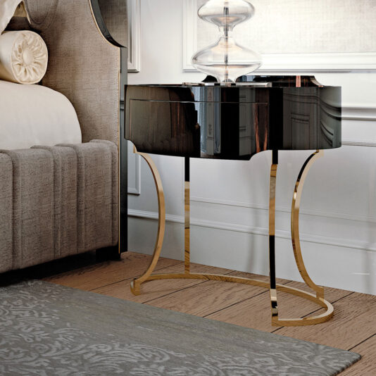 A modern bedroom showcases the Oval Italian Ebony Bedside Table, featuring sleek, curved golden legs next to a gray upholstered bed. A clear glass lamp sits on the nightstand, casting a soft light. The room includes a patterned area rug and boasts an elegant, minimalist decor.