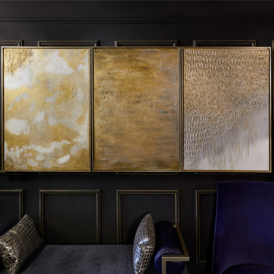 A sophisticated wall art display in a modern room showcases the "Set of Original Abstract Paintings," featuring diverse textures and presented in three large gold pieces. This artwork is elegantly complemented by a dark wall, a dark sofa with geometric patterned pillows, and a dark blue armchair, creating an overall refined atmosphere.