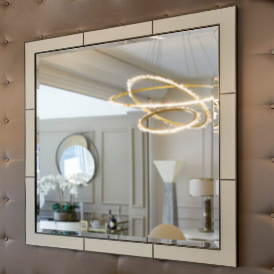 The Square Designer Leather Wall Mirror with its geometric frame is mounted on a padded wall. The mirror reflects an elegant, modern room adorned with crown molding, a circular chandelier featuring halo lights, an artwork piece, white orchids, and a contemporary table lamp.
