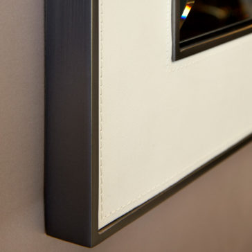Square Designer Leather Wall Mirror Close-up view of a picture frame displaying a corner of its edge. The frame has a sleek, black, metallic border and a beige, textured mat surrounding the image. The corner's angles and stitching emphasize the craftsmanship and design detail, reminiscent of the Square Designer Leather Wall Mirror.