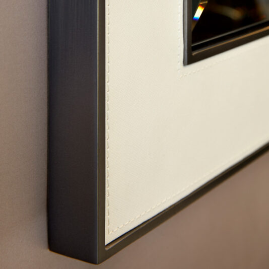 Close-up view of a picture frame displaying a corner of its edge. The frame has a sleek, black, metallic border and a beige, textured mat surrounding the image. The corner's angles and stitching emphasize the craftsmanship and design detail, reminiscent of the Square Designer Leather Wall Mirror.