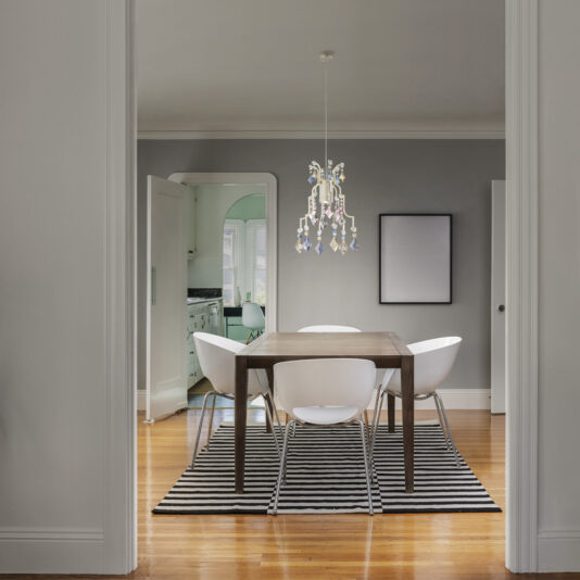 Traditional Crystal Pendant Chandelier In a contemporary dining area, a wooden table is encircled by four white chairs, all resting on a black and white striped rug. Suspended above the table is the Traditional Crystal Pendant Chandelier that adds an elegant touch to the space. The room features grey walls, an empty picture frame, and has a view of the kitchen in the background.