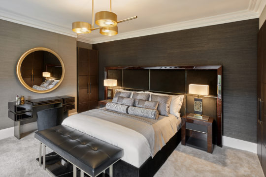 A modern bedroom featuring a large bed with grey linens and multiple pillows, flanked by nightstands with lamps. A cushioned bench sits at the foot of the bed. The room includes a dressing table with a round mirror and an upholstered chair. A sleek chandelier hangs from the ceiling.