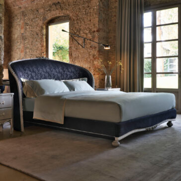 Classic Style Upholstered Winged Bed A cozy bedroom boasts a Classic Style Upholstered Winged Bed with a tall, curved headboard, adorned in light gray bedding. This elegant bed is set against a textured brick wall, accompanied by a large window dressed in sheer curtains that let in natural light. Above it, a modern wall lamp adds the perfect touch of illumination.