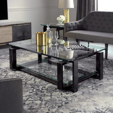 A modern living room featuring the Contemporary Glass Top Coffee Table with a sleek black frame, adorned with a gold vase and glassware. The space also includes a gray tufted sofa, a side table holding a lamp and flowers, and a patterned gray rug covering the floor. The overall ambiance of the room is both elegant and sophisticated.