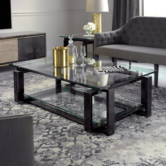 A modern living room featuring the Contemporary Glass Top Coffee Table with a sleek black frame, adorned with a gold vase and glassware. The space also includes a gray tufted sofa, a side table holding a lamp and flowers, and a patterned gray rug covering the floor. The overall ambiance of the room is both elegant and sophisticated.
