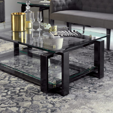 A contemporary living room features the Contemporary Glass Top Coffee Table, showcasing a sleek black metal frame. The table is adorned with a gold ice bucket, two empty champagne flutes, a clear carafe, and a magazine. The floor is covered with a gray patterned rug, while a gray tufted sofa sits in the background.