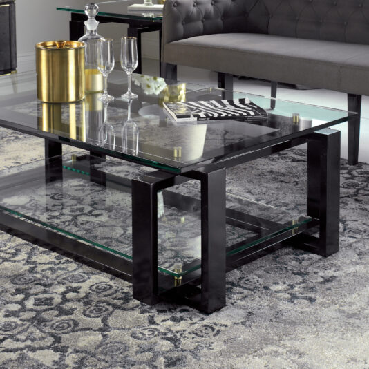 Contemporary Glass Top Coffee Table A contemporary living room features the Contemporary Glass Top Coffee Table, showcasing a sleek black metal frame. The table is adorned with a gold ice bucket, two empty champagne flutes, a clear carafe, and a magazine. The floor is covered with a gray patterned rug, while a gray tufted sofa sits in the background.