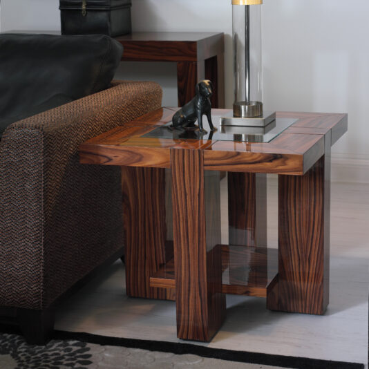 A modern living room features a Contemporary Square End Table With Glass Top beside a cushioned armchair. The table holds a sleek black statue and a contemporary lamp with a transparent base, all set on a light-colored floor and patterned rug.