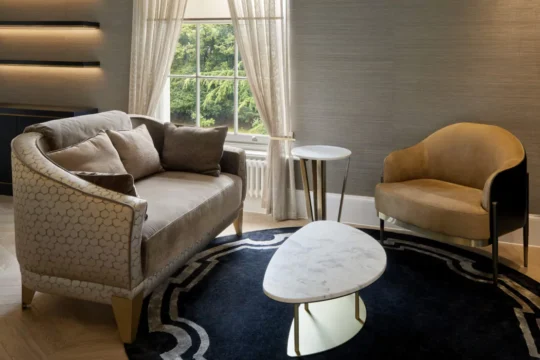 A stylish living room with a gray couch and beige armchair on a dark circular rug, featuring a marble-topped coffee table with gold accents. Natural light filters through a window with sheer curtains, illuminating the modern decor.