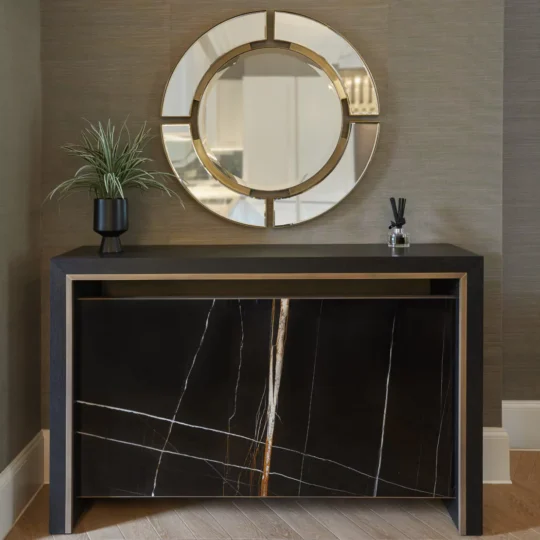 A modern entryway features a black console table with a reflective marble design. Above it hangs a round mirror with gold accents. A small plant and a reed diffuser sit on the console. The walls are textured, and the floor is wooden.