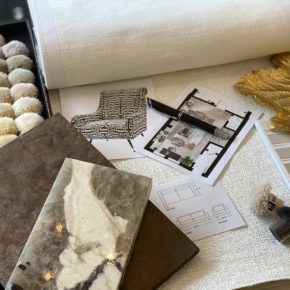 A tabletop with assorted interior design materials: a color swatch book, fabric samples, architectural plans, a pen, brown textured boards, a marbled tile, and a gold leaf decor piece. The setup suggests planning for a home or room design. Learn Interior Design with us to make your vision come to life.