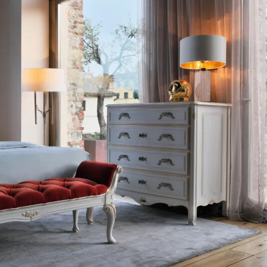 Tall Louis Style Chest Of Drawers A cozy bedroom featuring a white dresser with ornate handles, topped with a lamp and a golden decorative item. Beside it stands proudly the Tall Louis Style Chest Of Drawers. There's also a red cushioned bench at the foot of the bed, next to large windows with sheer curtains, allowing soft natural light in.