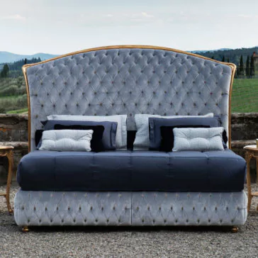 Button Upholstered Classic Style Bed The Button Upholstered Classic Style Bed is a luxurious piece with an elegantly tufted velvet headboard in a light blue shade, adorned with gold accents. This exquisite bed features multiple pillows in black, white, and checkered patterns and is beautifully set outdoors against a backdrop of rolling hills and greenery.