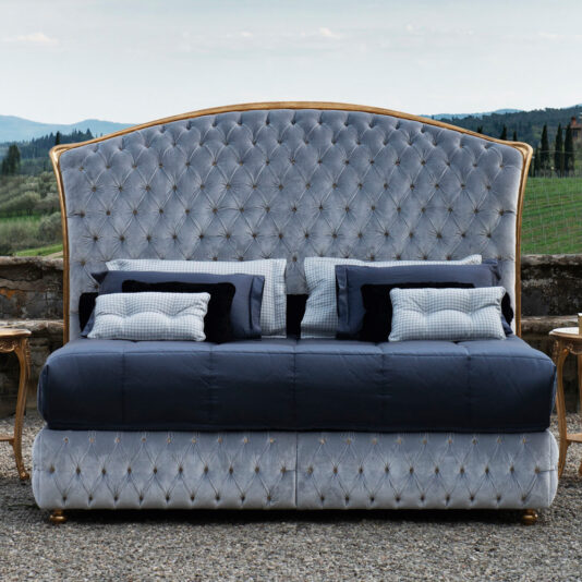 The Button Upholstered Classic Style Bed is a luxurious piece with an elegantly tufted velvet headboard in a light blue shade, adorned with gold accents. This exquisite bed features multiple pillows in black, white, and checkered patterns and is beautifully set outdoors against a backdrop of rolling hills and greenery.