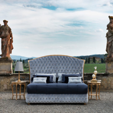 Button Upholstered Classic Style Bed A Button Upholstered Classic Style Bed with a tufted, plush headboard and navy bedding is flanked by two side tables, each adorned with an ornate lamp. The bed is set against a backdrop featuring a scenic rural landscape with vineyards, statues, and a partly cloudy sky.
