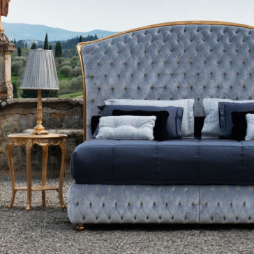 Button Upholstered Classic Style Bed A Button Upholstered Classic Style Bed with a tufted, curved headboard is elegantly dressed in dark blue and gray bedding and stands outdoors on a gravel surface. Next to the bed is a stylish gold side table with a matching lamp. In the background, a scenic landscape with greenery and hills unfolds.