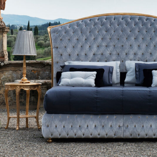 A Button Upholstered Classic Style Bed with a tufted, curved headboard is elegantly dressed in dark blue and gray bedding and stands outdoors on a gravel surface. Next to the bed is a stylish gold side table with a matching lamp. In the background, a scenic landscape with greenery and hills unfolds.