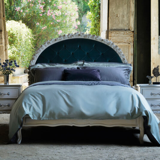 A cozy bedroom featuring the Carved Rose Button Upholstered Bed with a tufted, arched headboard. The bed is adorned with blue and gray bedding. Two nightstands with drawers flank either side, each showcasing a decorative item. Rustic wooden doors open to reveal lush greenery outside.