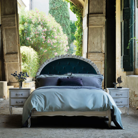 Carved Rose Button Upholstered Bed A luxurious bedroom showcases the Carved Rose Button Upholstered Bed, featuring a large, tufted headboard adorned with intricate carvings. The bed is dressed with plush velvet and satin pillows and an elegant blue duvet. On either side of the bed are white nightstands topped with lamps and vases of flowers. Through the open rustic wooden doors, lush greenery is visible in the background.