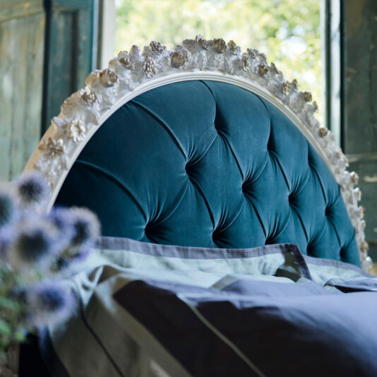 Carved Rose Button Upholstered Bed A plush, teal tufted headboard with ornate white floral details at the top is positioned in a cozy bedroom. The Carved Rose Button Upholstered Bed is adorned with neatly arranged gray and white pillows and blankets. In the foreground, there is a blurred bouquet of purple flowers.