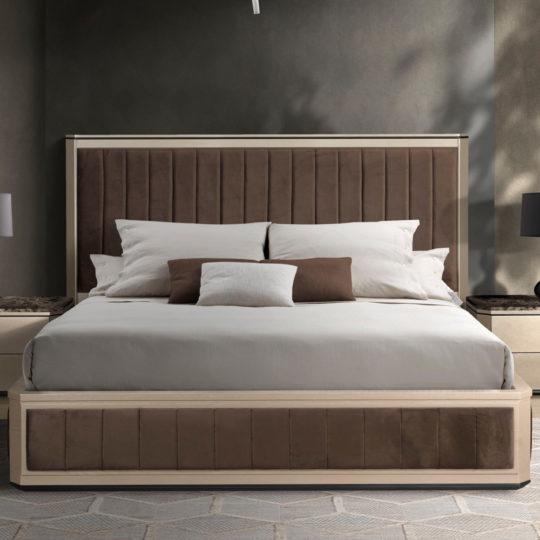 A Chic Contemporary Upholstered Bed features a brown headboard and matching footboard, adorned with a neutral-toned bedspread. The bedding set includes white pillows and cushions with a central brown accent pillow. Nightstands with lamps are positioned on either side of the bed against a dark wall.