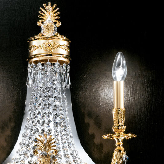 Classic Crystal Empire Style Wall Light A close-up of an ornate chandelier part and a candle-style light fixture. The Classic Crystal Empire Style Wall Light features classic gold accents, cascading crystal beads, a gold base with intricate detailing, and a flame-shaped bulb. The background is black.
